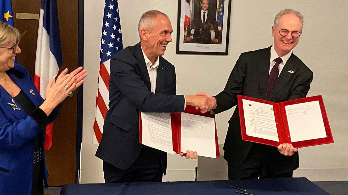 Two people are shaking hands and smiling while holding copies of the agreement. A third person is standing nearby and clapping.